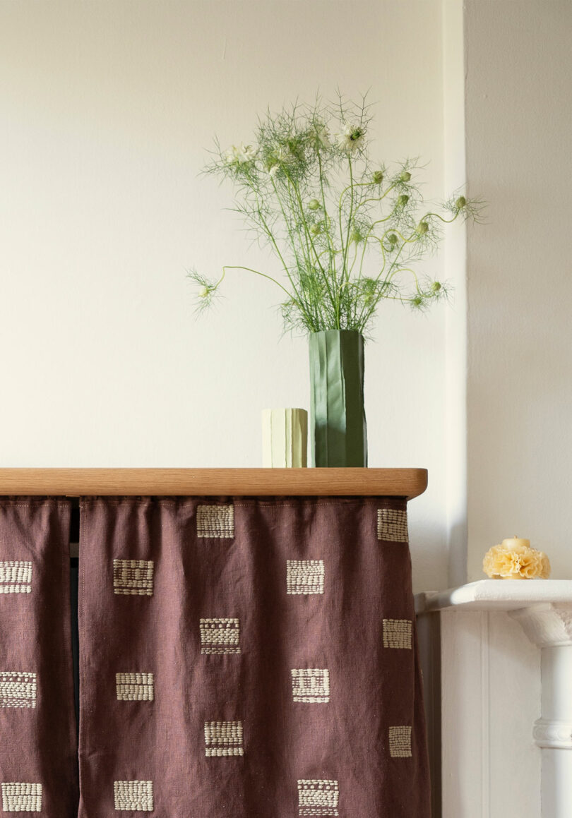 A green vase with wispy greenery and a small book sit on a wooden surface with a patterned brown fabric draped below. A yellow flower rests on a nearby ledge.