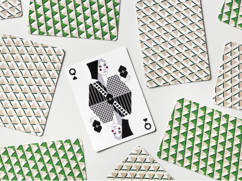 A queen of clubs playing card with a modern geometric design is face up among several face-down cards with green and beige patterned backs.