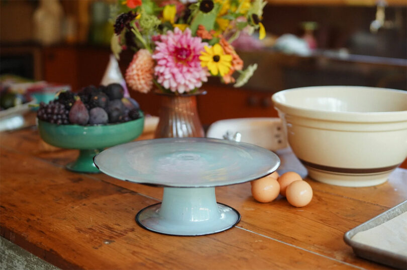 A cake stand, three eggs, a bowl of fruit, a vase of flowers, a large mixing bowl, and a colander sit on a wooden kitchen counter—perfect inspiration for thoughtful Valentine’s Day gifts.