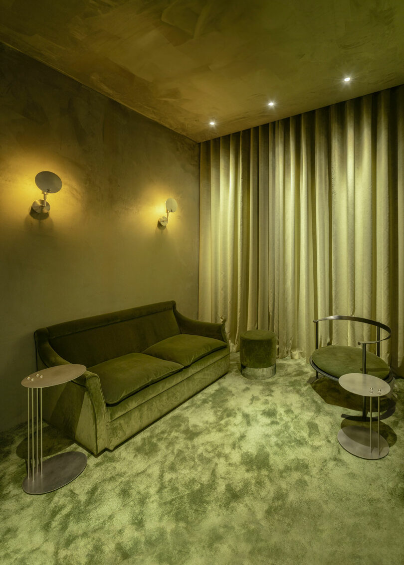 A modern lounge with green walls, carpet, velvet sofa, round ottoman, metal side tables, round glass coffee table, and floor-to-ceiling drapes under recessed lighting.