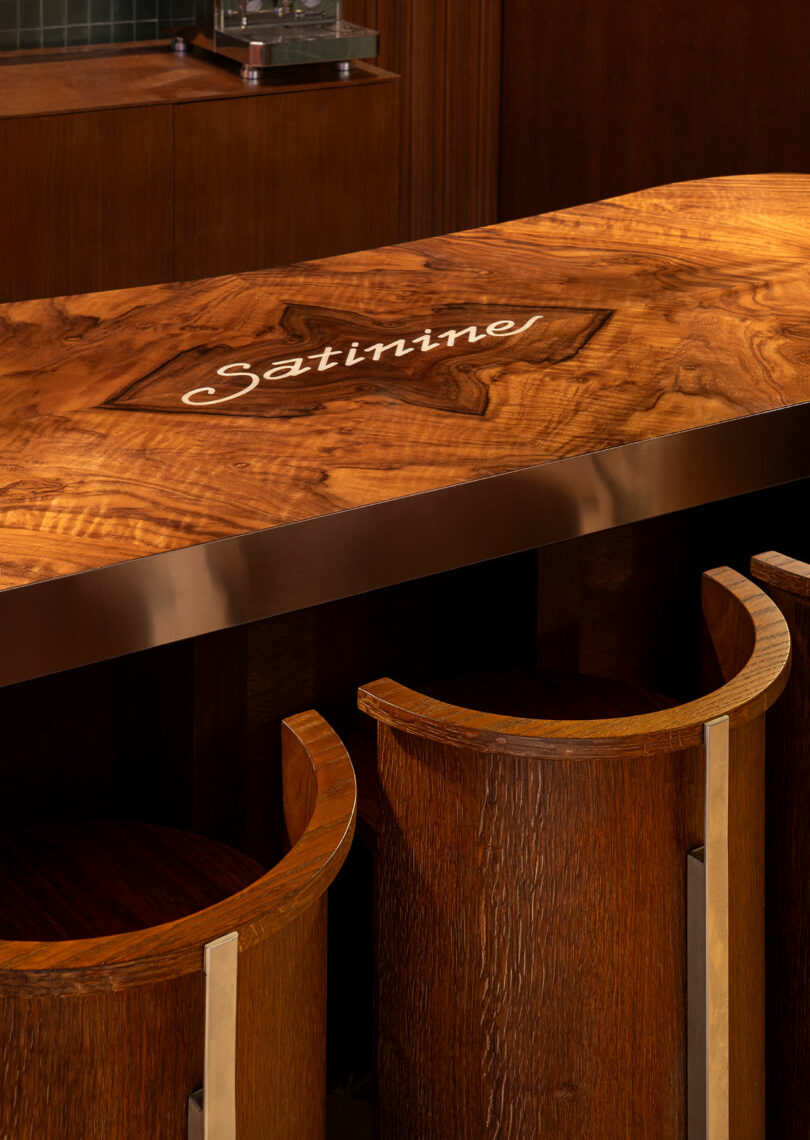 A polished wooden bar counter with the word "Satinine" engraved on the surface, accompanied by three curved wooden bar stools.