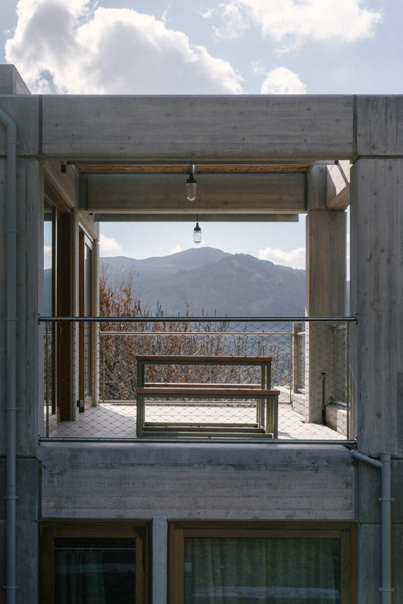 A concrete balcony by Meraki Studios, with a table and bench, overlooks distant mountains beneath a partly cloudy sky. A hanging lightbulb is centered above the serene space.