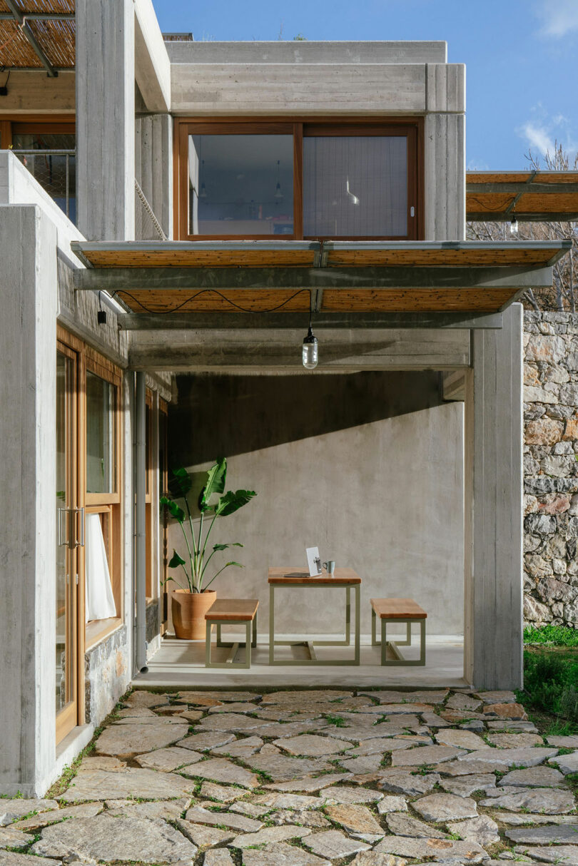 Modern concrete patio by Meraki Studios with a wooden table, benches, potted plant, overhead metal awning, and large windows; stone-paved ground and rocky wall complete the stylish look.