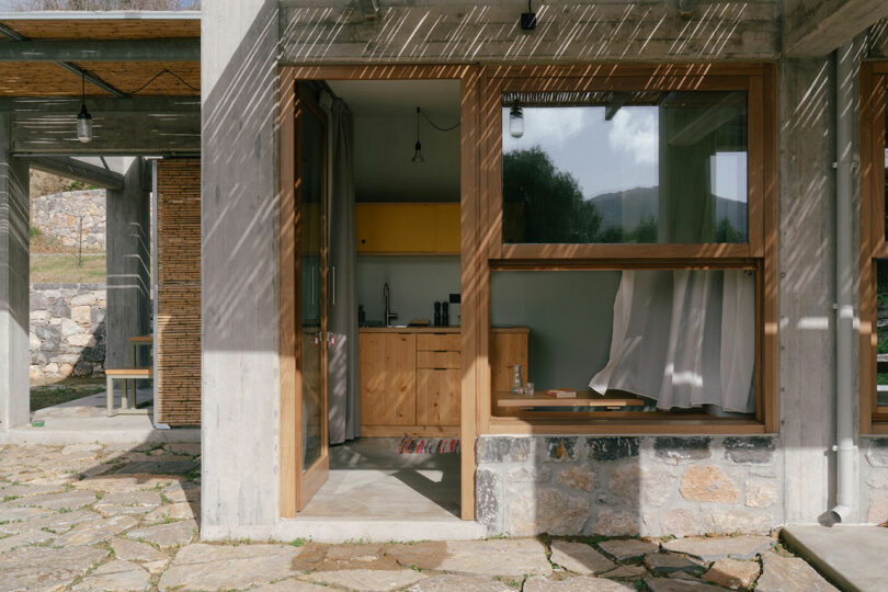 A modern home by Meraki Studios with concrete walls and large wooden-framed windows; sunlight creates patterns on the stone patio, and a small kitchen is visible inside.