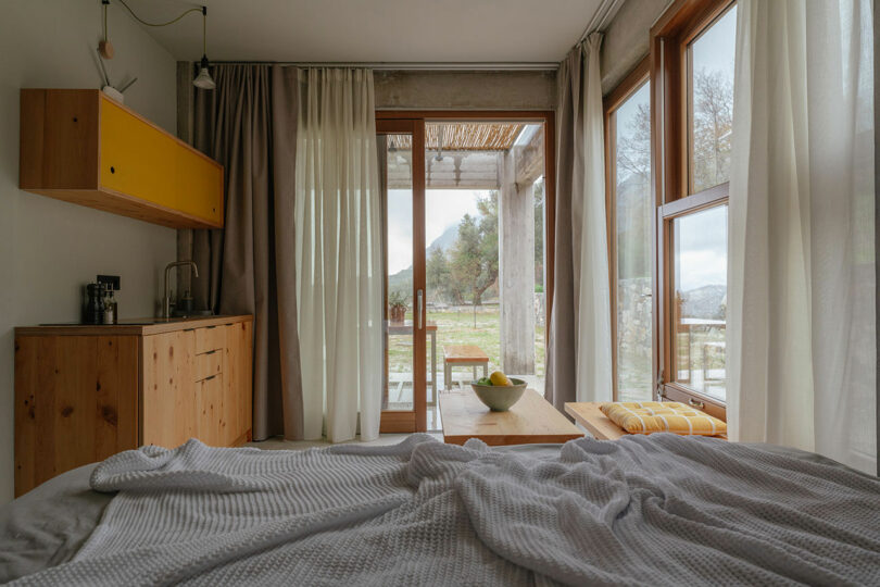 A cozy Meraki Studios bedroom with an unmade bed, wooden kitchenette, and large windows draped in sheer curtains overlooking a patio and lush green outdoor landscape.