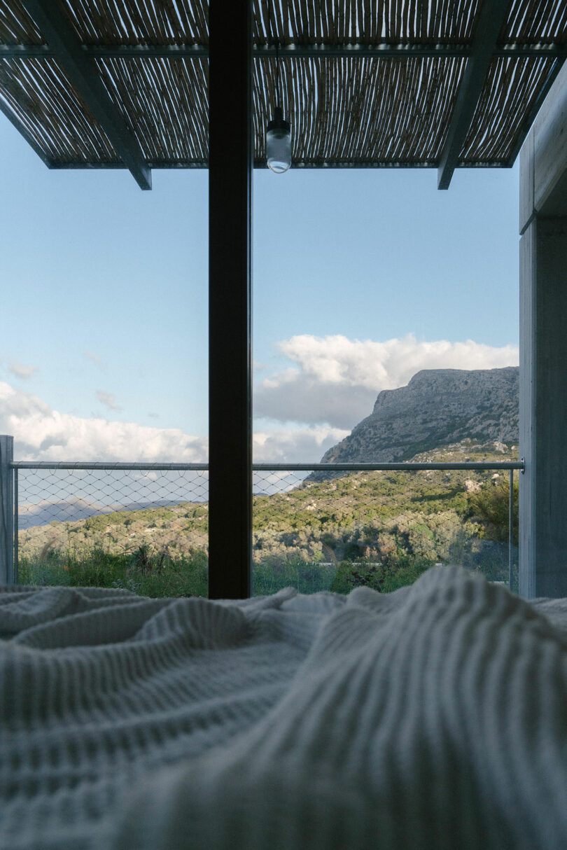 View from a bed looking out through a large window at a mountain landscape, with a slatted pergola overhead and a metal railing on the balcony outside—crafted by Meraki Studios.