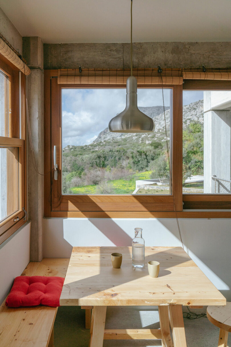 A small wooden dining table by Meraki Studios, adorned with a glass bottle and two cups, sits by large windows showcasing a mountain view, bathed in natural light with a hanging pendant lamp above.