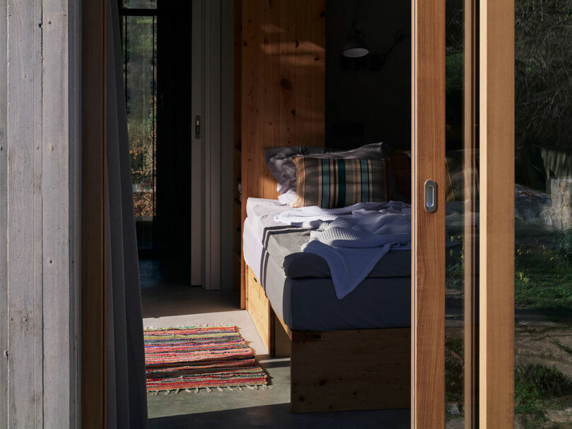 A sunlit bedroom by Meraki Studios features an unmade bed with a wooden frame, plaid pillows, and a colorful rug, all viewed through an open sliding glass door.