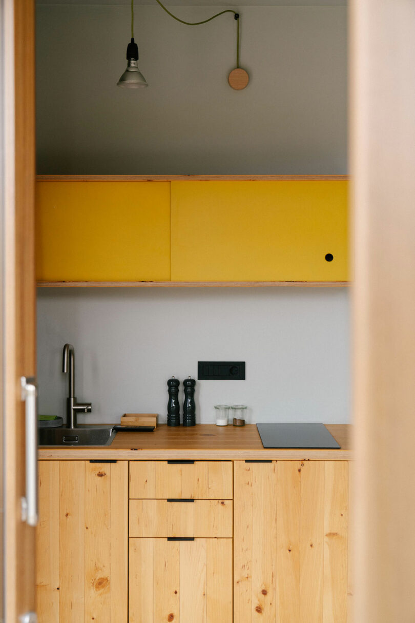 A compact kitchen by Meraki Studios features light wooden cabinets, a yellow wall-mounted cupboard, a sink, a stovetop, and stylish kitchen accessories on the counter.