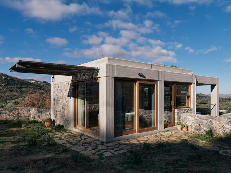 Modern single-story house by Meraki Studios with large glass windows, concrete and stone exterior, set on a stone patio in a rural landscape under a partly cloudy sky.