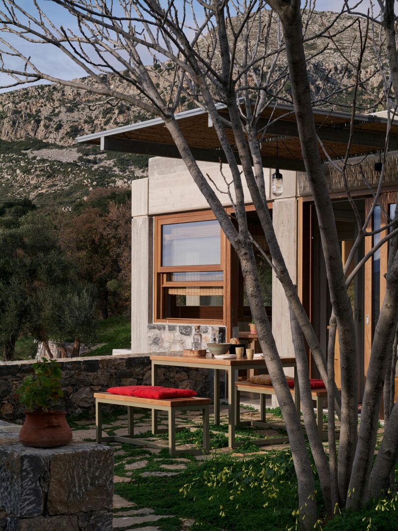 A modern house by Meraki Studios with large windows and a small outdoor seating area featuring red cushions and a table, set against a rocky hillside and surrounded by trees.