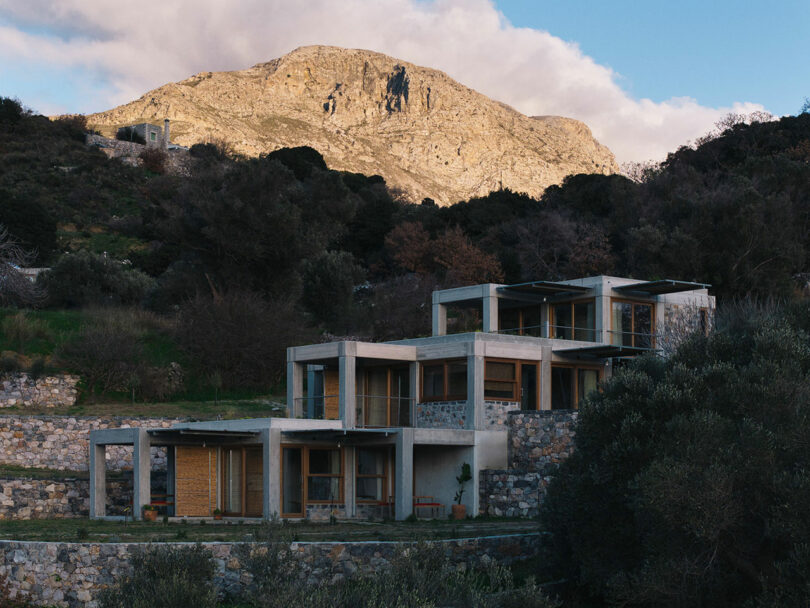 Modern concrete house with large windows and stone walls, designed by Meraki Studios, situated on a hillside with a rugged mountain in the background under a partly cloudy sky.