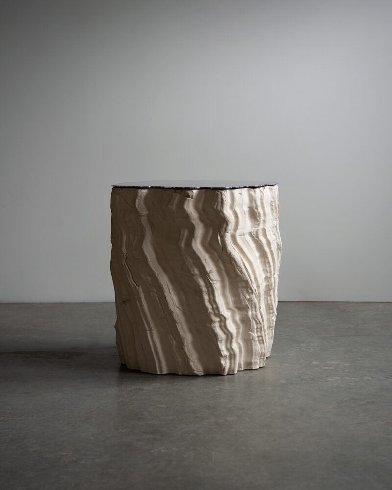 A cylindrical, beige sculptural object with a textured, striated surface stands on a smooth concrete floor against a plain gray wall.