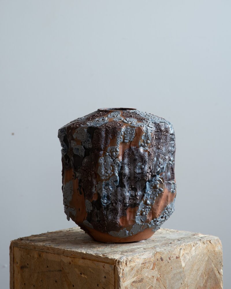 A ceramic vase with a rough, textured surface featuring dark, metallic and ashy blue glazes, displayed on a plywood pedestal against a plain background.