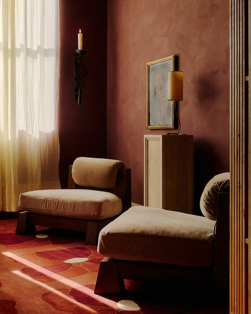 Two cushioned chairs sit in a dimly lit room with textured red walls, a tall window with sheer curtains, abstract art, a lamp, and a wall-mounted candleholder.