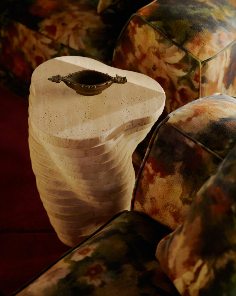 A sculpted stone side table with layered, organic shapes holds a small decorative dish, placed between floral patterned upholstered chairs.