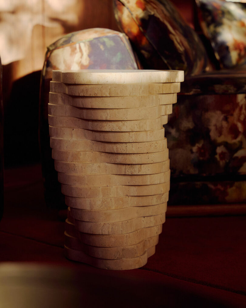 A sculptural, layered beige stone side table with a twisting design sits on a reddish floor next to a floral-patterned chair.