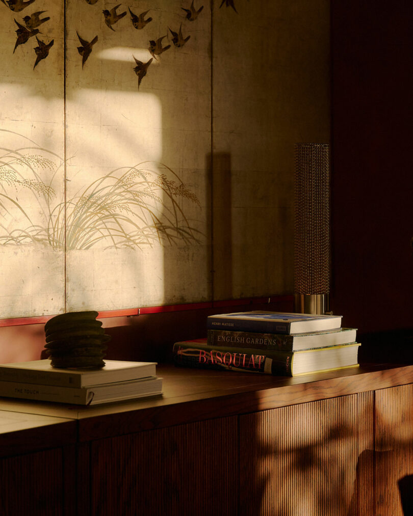 A stack of books and a decorative object on a wooden sideboard, with sunlight casting shadows and a bird-themed folding screen in the background.