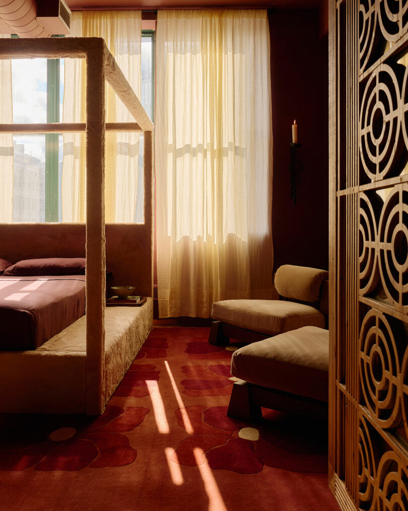 A sunlit bedroom with sheer curtains, a canopy bed, two cushioned chairs, and a carpet with circular patterns next to a decorative wooden screen.