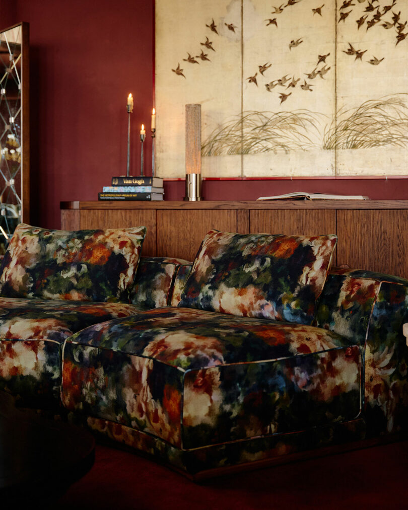 A floral-patterned sofa sits in front of a wooden sideboard with candles, books, and a decorative screen featuring flying birds and grass, set against a deep red wall.