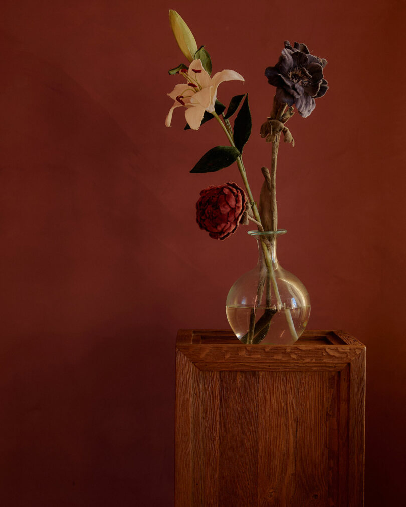 A clear glass vase with three flowers—one white, one red, and one dark purple—stands on a wooden pedestal against a reddish-brown background.