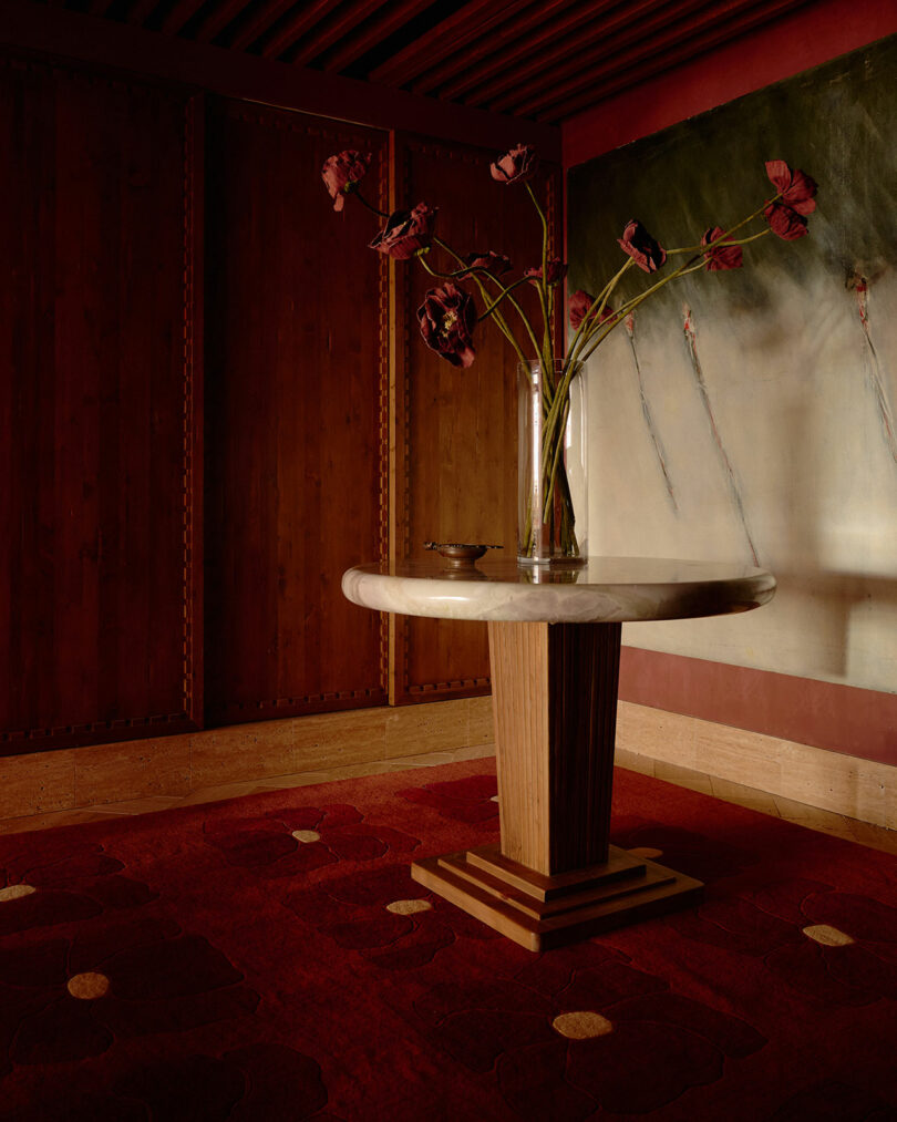 A round marble-top table with a wooden base stands on a red carpet, holding a tall glass vase with long-stemmed dark red flowers, against wood-paneled walls and a large mural.