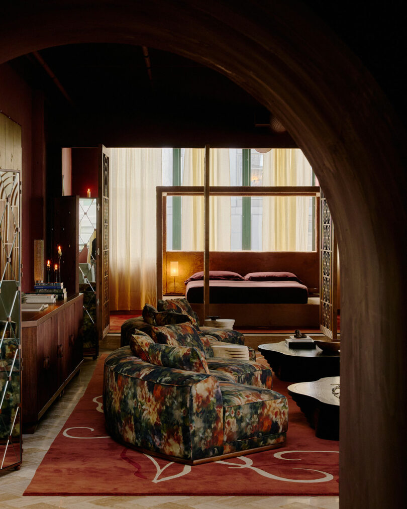 A dimly lit room with floral-patterned sofas, a red rug, and a canopy bed in the background, viewed through an arched doorway.