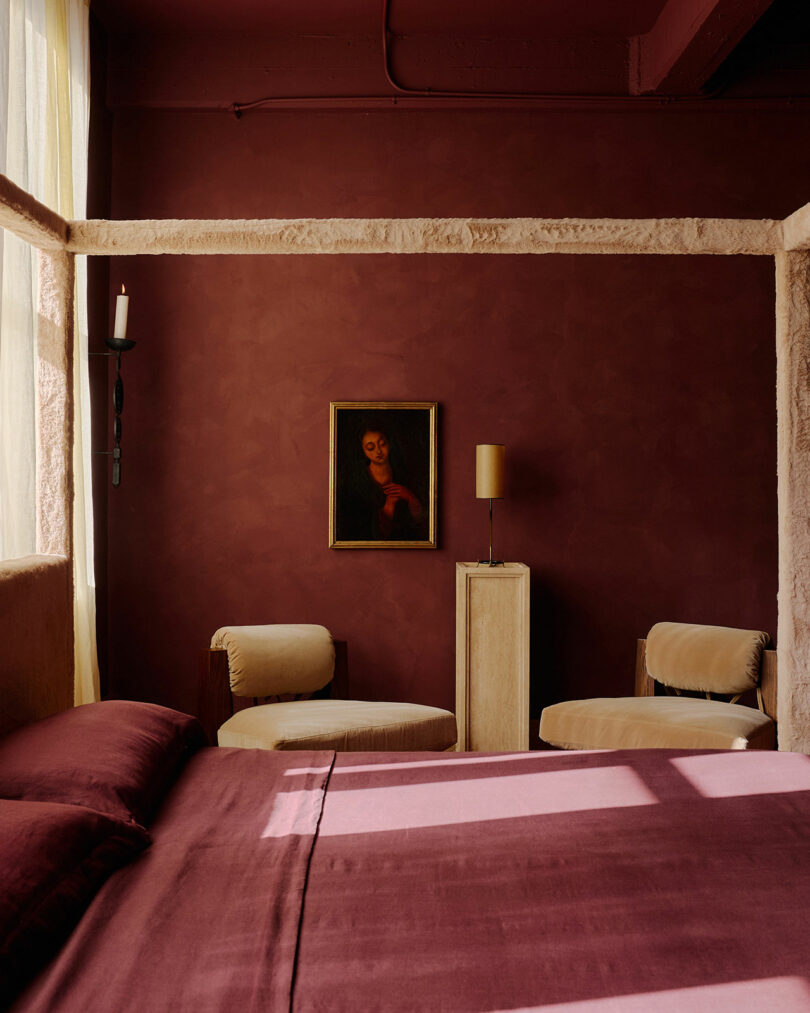 A bedroom with burgundy walls and bedding, two beige chairs, a framed portrait, a small lamp on a pedestal, and sunlight streaming through a window.