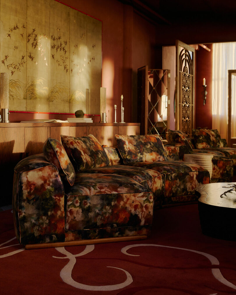 A living room with patterned floral sofas, a wooden coffee table, red walls, a decorative screen, and a large wall art piece featuring birds. Natural light enters through sheer curtains.