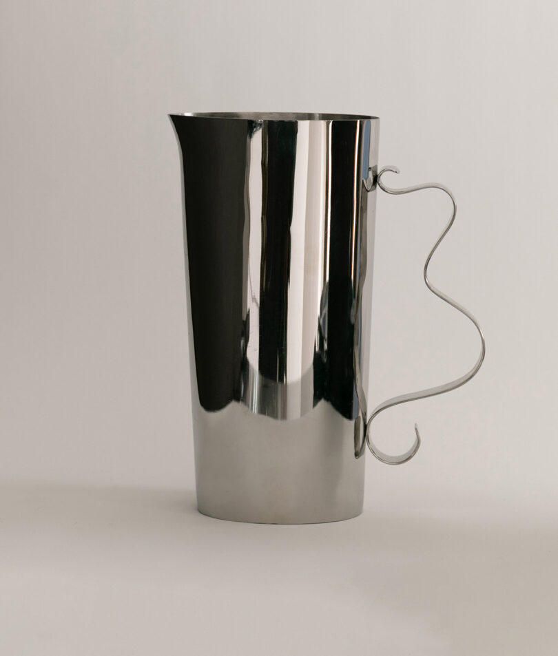 A tall, reflective metal pitcher with a curved, decorative handle is placed on a light surface against a plain background.