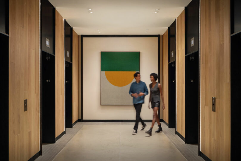 Two people walk through a modern hallway lined with elevator doors, featuring a geometric artwork at the end—an interior space designed by Morris Adjmi Architects.