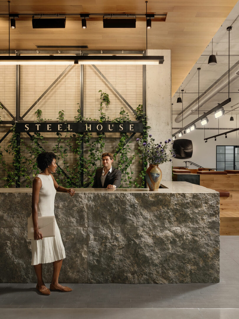 A woman in a white dress talks to a man at a stone reception desk in a modern lobby designed by Morris Adjmi Architects, with "Steel House" signage and green plants behind the counter.