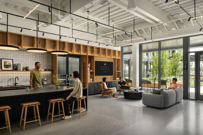 Modern office lounge designed by Morris Adjmi Architects, featuring people conversing, a kitchenette with bar stools, large windows, and a "Steel House" sign on the wall.