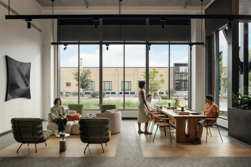 Modern office lounge designed by Morris Adjmi Architects, featuring large windows, four people interacting on sofas and chairs, with a long wooden table and lush plants enhancing the space.
