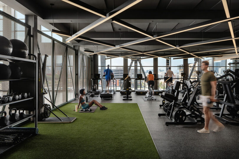 Modern gym designed by Morris Adjmi Architects with large windows, several people using cardio and strength equipment, and one person sitting on artificial turf next to gym gear.