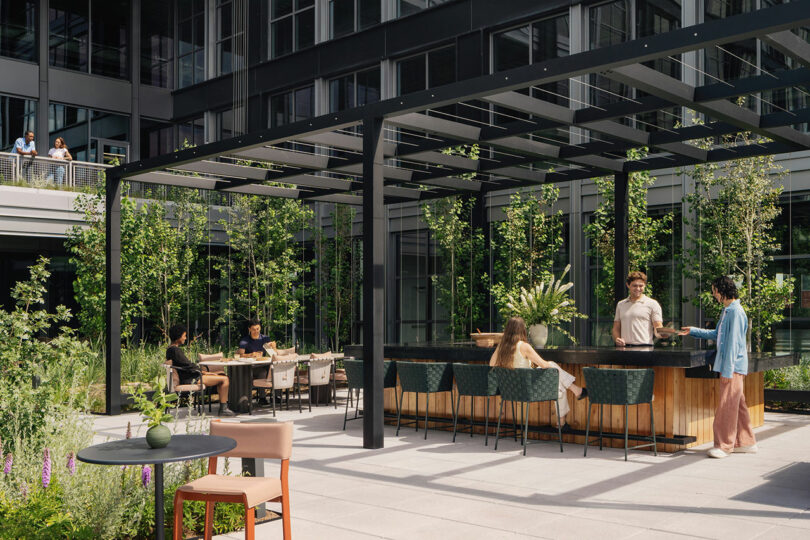 People sit and stand around an outdoor bar and tables in a modern courtyard with greenery, shaded by a black pergola designed by Morris Adjmi Architects, adjacent to a glass office building.