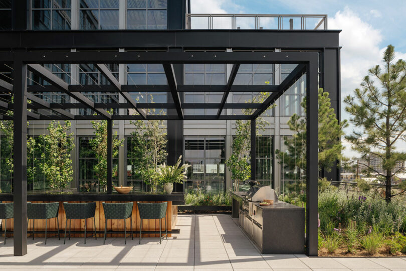 Outdoor rooftop kitchen designed by Morris Adjmi Architects, featuring a long dining table, chairs, built-in grill, ample counter space, and a pergola structure surrounded by greenery with a modern building in the background.