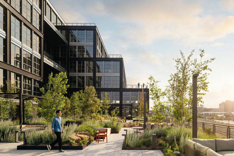A person walks along a landscaped rooftop terrace with seating areas and greenery, designed by Morris Adjmi Architects, beside a modern glass office building under a partly cloudy sky.