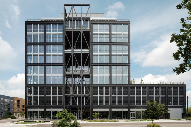 A modern, multi-story office building designed by Morris Adjmi Architects features a black steel frame and large glass windows, viewed from the front on a partly cloudy day.