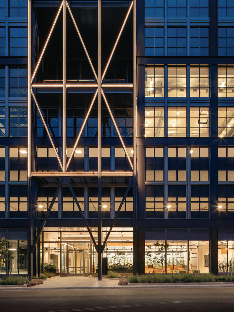 Modern building facade at dusk with illuminated linear lights forming geometric patterns, reflecting the signature style of Morris Adjmi Architects; glass windows reveal lit interior spaces and main entrance at street level.