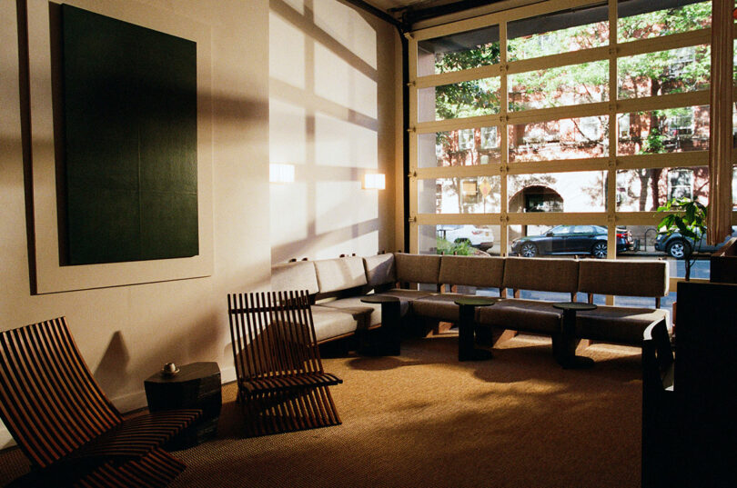 Modern waiting area with wooden chairs, cushioned benches, a large abstract painting, and a glass garage-style door letting in natural light.