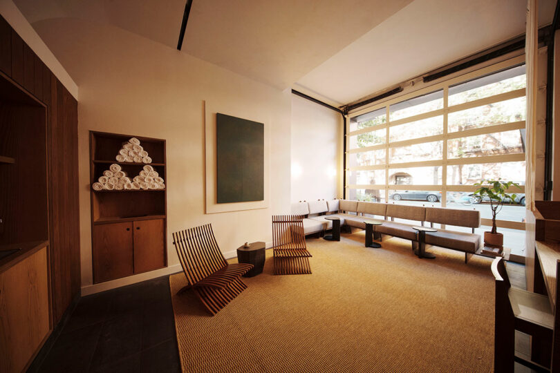 Minimalist lounge with wooden slat chairs, a long bench, stacked towels on shelves, large abstract wall art, and floor-to-ceiling windows showing a parked car outside.