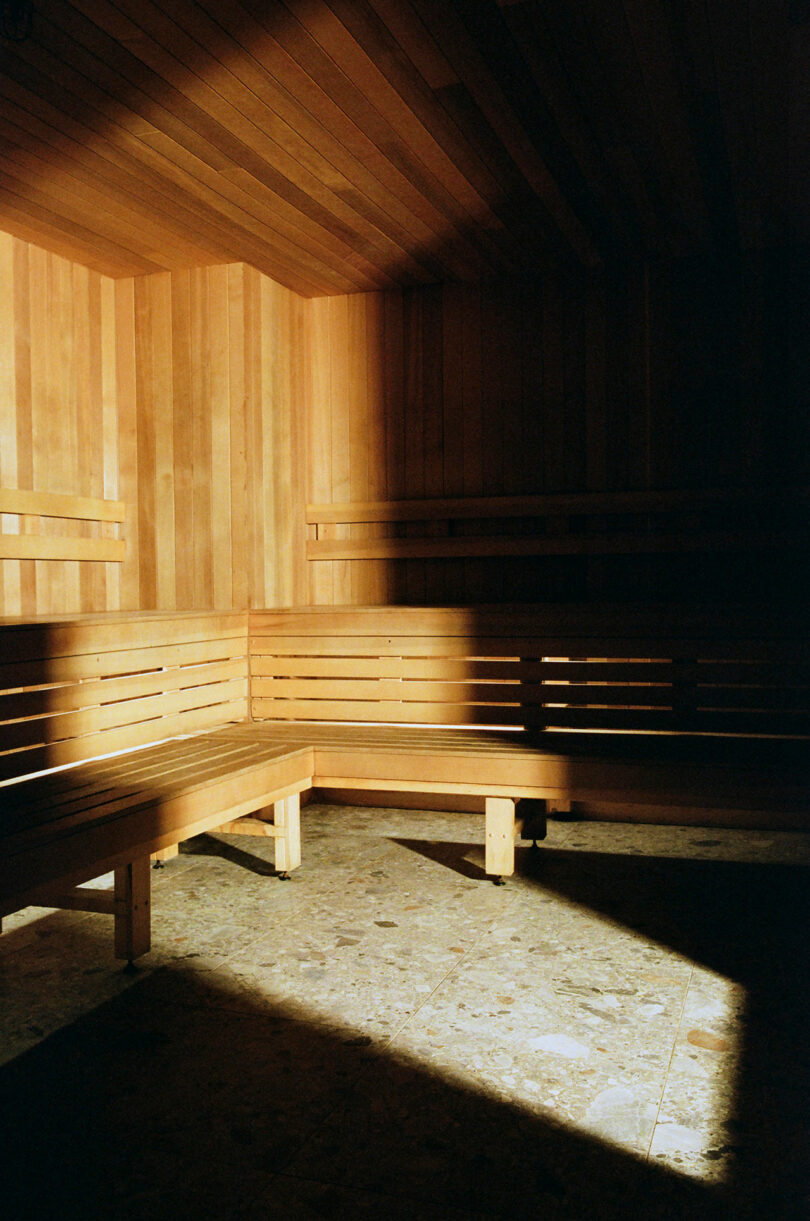 Empty wooden sauna with built-in benches and sunlight casting shadows on the tiled floor and walls.