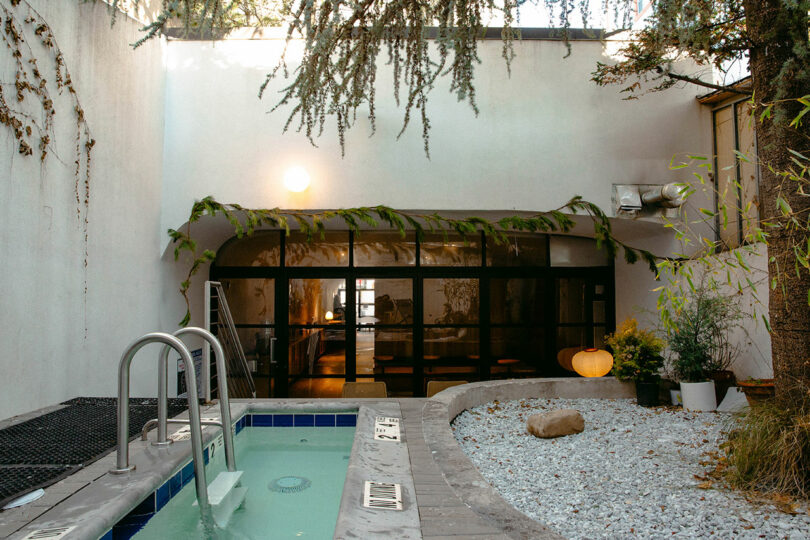 Outdoor spa area with a hot tub, gravel garden, potted plants, and a glass door leading to an indoor space. A tree and hanging branches are visible in the enclosed courtyard.