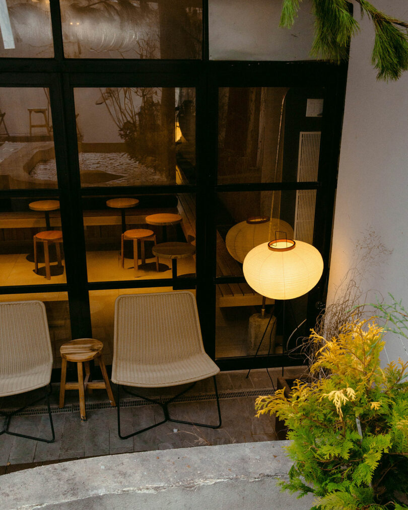Outdoor patio with two beige chairs, a small wooden stool, and a round paper lantern next to large glass doors revealing an indoor seating area with stools and warm lighting.