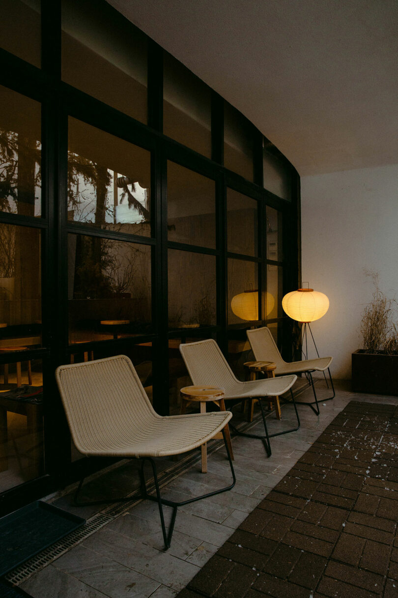 Three wicker lounge chairs with small wooden tables are arranged on a tiled patio, beside a large glass window and a glowing floor lamp.