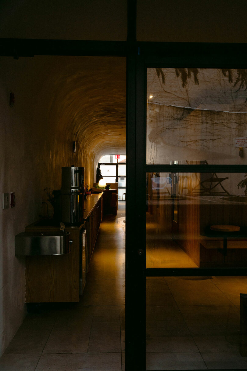 A dimly lit interior space with a curved ceiling, metal sink, counter, and seating area, viewed through a glass door.