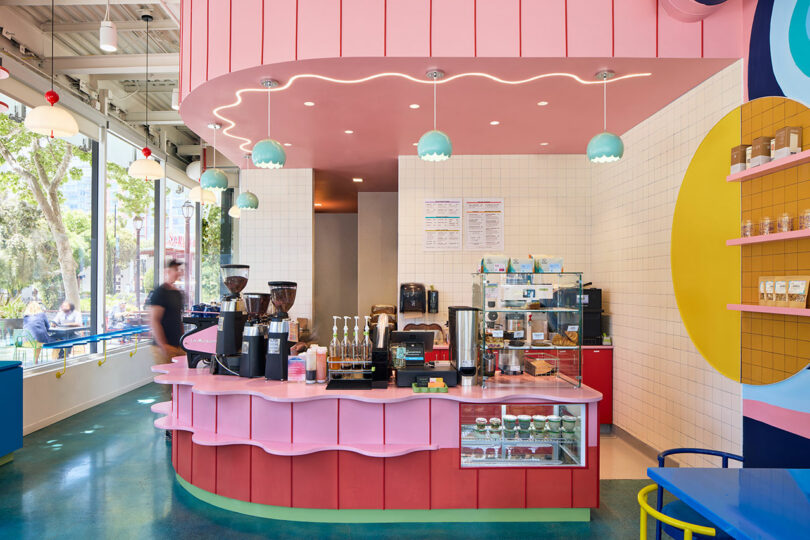 A brightly colored modern café interior with a pink counter, coffee machines, pastry display, and large windows showing trees outside.
