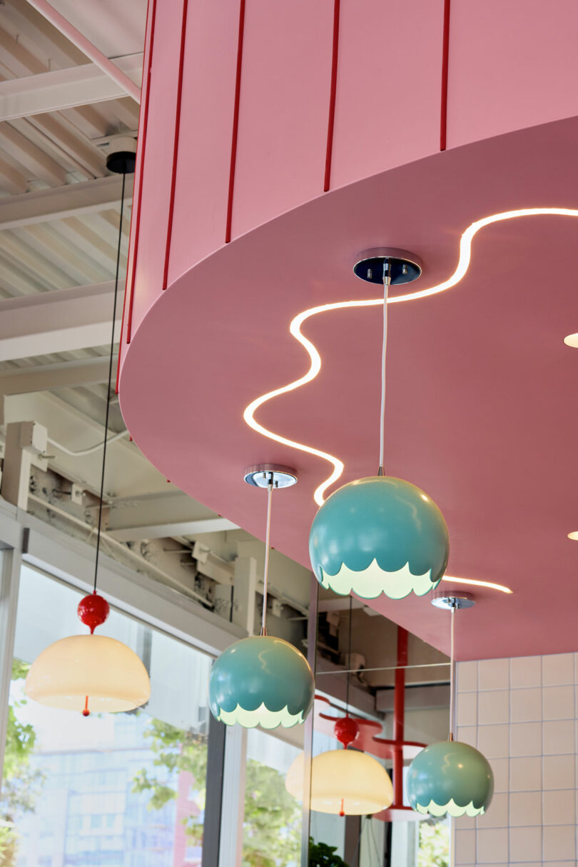 A pink ceiling with wavy recessed lighting and blue pendant lights with scalloped edges hangs above a modern, brightly lit interior.