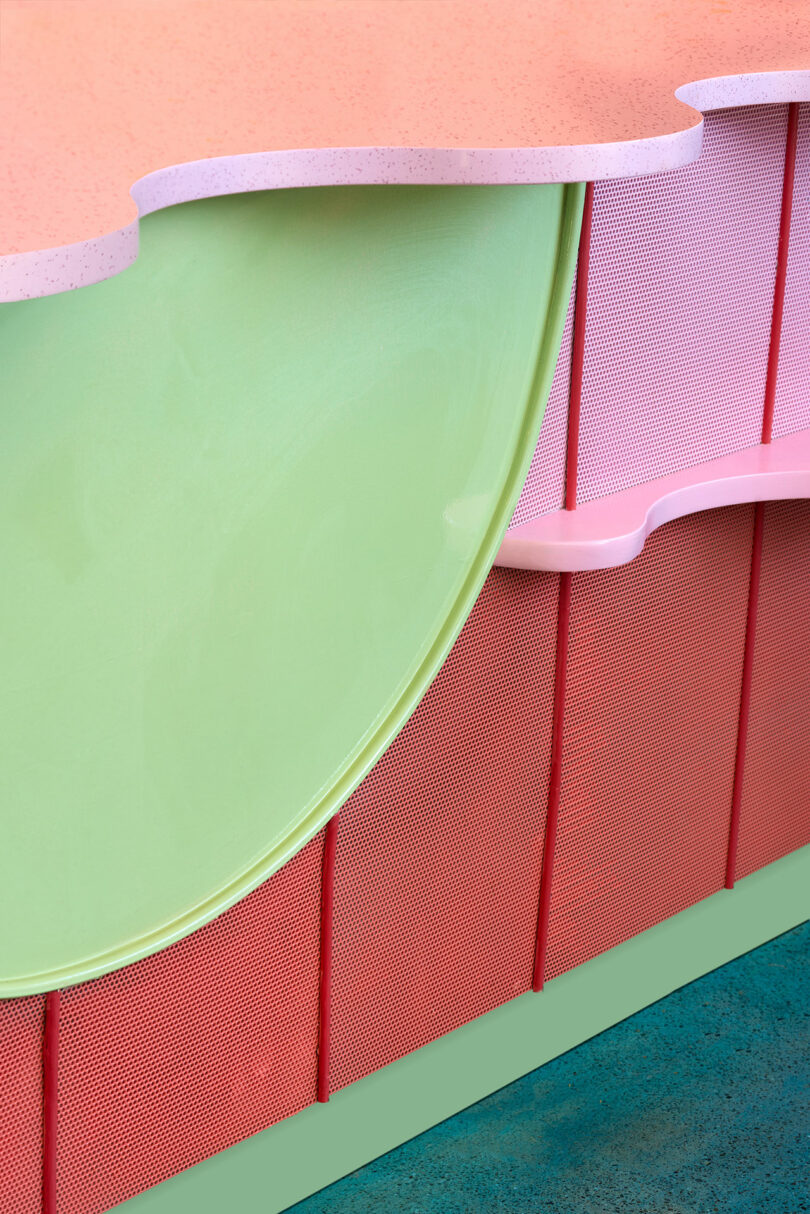Close-up of a modern furniture piece with wavy pink edging, a smooth green circular surface, and red textured panels, set on a blue floor.
